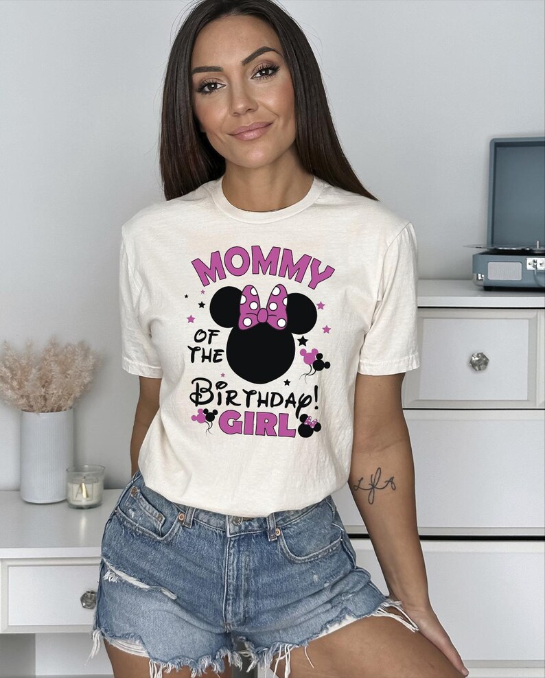 May include: Cream-coloured t-shirt with the text "MOMMY OF THE BIRTHDAY GIRL!" in pink and black. The design includes a Minnie Mouse silhouette with a pink polka-dot bow. The shirt is paired with denim shorts.