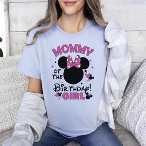 May include: Pale blue t-shirt featuring the text "MOMMY OF THE BIRTHDAY GIRL" in pink and black. The design includes a black mouse head silhouette with a pink bow and mouse silhouettes. The shirt has short sleeves.