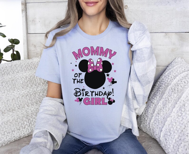 May include: Pale blue t-shirt featuring the text "MOMMY OF THE BIRTHDAY GIRL" in pink and black. The design includes a black mouse head silhouette with a pink bow and mouse silhouettes. The shirt has short sleeves.