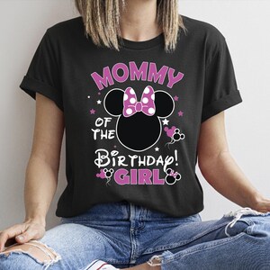 May include: Black t-shirt with the text "MOMMY OF THE BIRTHDAY GIRL!" in pink and white. The design includes a Minnie Mouse silhouette with a polka dot bow. The shirt is worn by a person wearing blue jeans.
