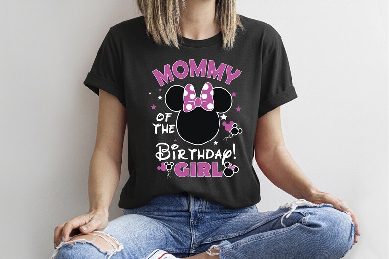 May include: Black t-shirt with the text "MOMMY OF THE BIRTHDAY GIRL!" in pink and white. The design includes a Minnie Mouse silhouette with a polka dot bow. The shirt is worn by a person wearing blue jeans.