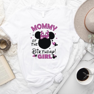 May include: White t-shirt with a Minnie Mouse design. The design features a black Minnie Mouse head with a pink bow, the text "Mommy of the Birthday Girl" in pink and black, and star accents. The shirt is displayed with a hat, coffee, and a book.