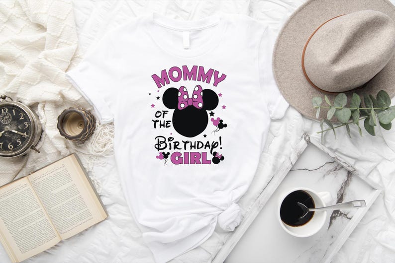 May include: White t-shirt with a Minnie Mouse design. The design features a black Minnie Mouse head with a pink bow, the text "Mommy of the Birthday Girl" in pink and black, and star accents. The shirt is displayed with a hat, coffee, and a book.
