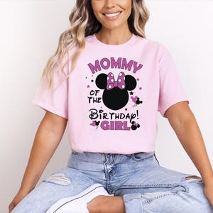 May include: A pale pink t-shirt featuring the text "MOMMY OF THE BIRTHDAY GIRL" in purple and black. The design includes a black Minnie Mouse head with a purple bow and stars. The shirt is short-sleeved.