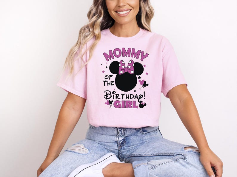 May include: A pale pink t-shirt featuring the text "MOMMY OF THE BIRTHDAY GIRL" in purple and black. The design includes a black Minnie Mouse head with a purple bow and stars. The shirt is short-sleeved.