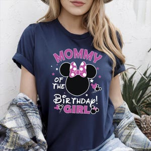 May include: Navy blue t-shirt featuring the text "MOMMY OF THE BIRTHDAY GIRL!" in pink and white. The design includes a black Minnie Mouse silhouette with a pink polka dot bow and pink accents. A casual, festive top.