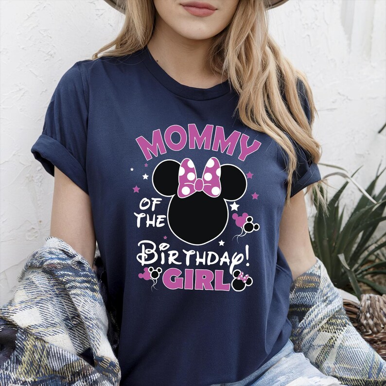 May include: Navy blue t-shirt featuring the text "MOMMY OF THE BIRTHDAY GIRL!" in pink and white. The design includes a black Minnie Mouse silhouette with a pink polka dot bow and pink accents. A casual, festive top.