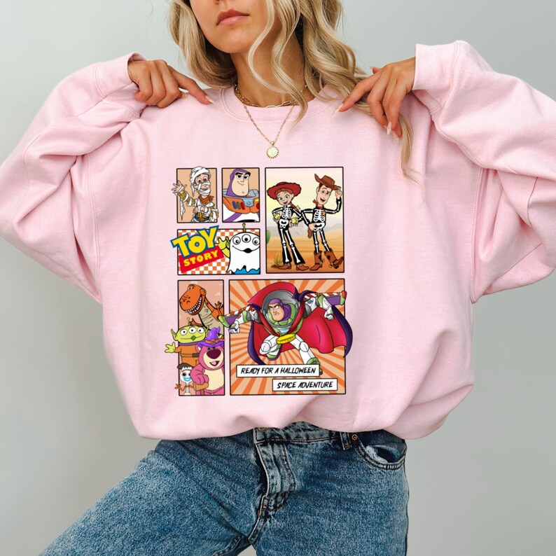 May include: Light pink sweatshirt featuring a comic strip design with characters from the Toy Story movie. The design includes Buzz Lightyear, Woody, and other characters. The text "READY FOR A HALLOWEEN SPACE ADVENTURE" is also visible.