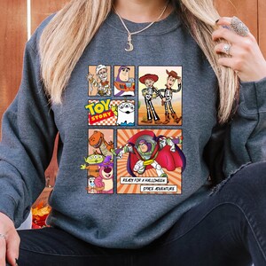 May include: A dark gray sweatshirt featuring a colorful graphic of Toy Story characters in a comic book style. The design includes Buzz Lightyear, Woody, and other characters, with the text "READY FOR A HALLOWEEN SPACE ADVENTURE."