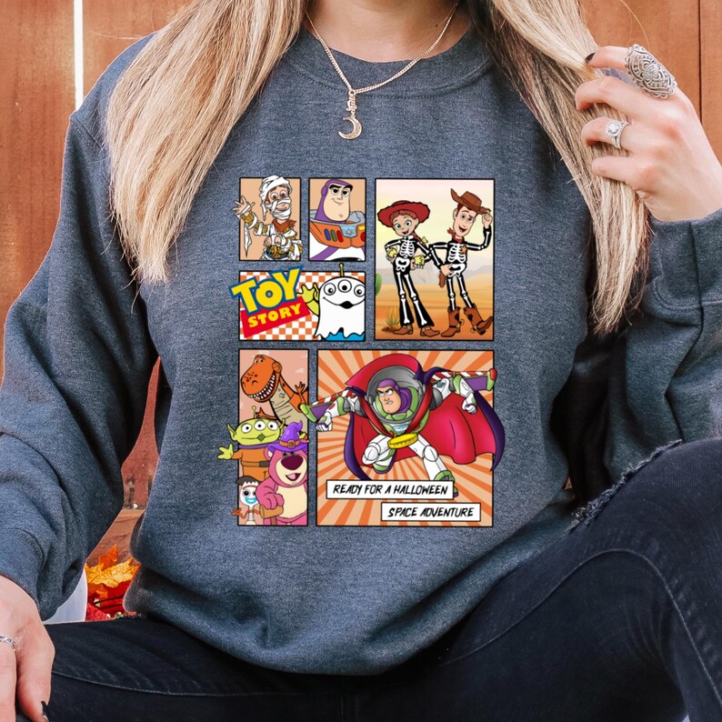 May include: A dark gray sweatshirt featuring a colorful graphic of Toy Story characters in a comic book style. The design includes Buzz Lightyear, Woody, and other characters, with the text "READY FOR A HALLOWEEN SPACE ADVENTURE."