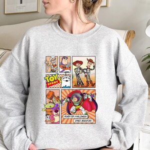 May include: A light gray sweatshirt featuring a comic strip design with characters from the movie "Toy Story." The design includes Buzz Lightyear, Woody, and other characters. Text on the design reads "TOY STORY", "READY FOR A HALLOWEEN SPACE ADVENTURE."
