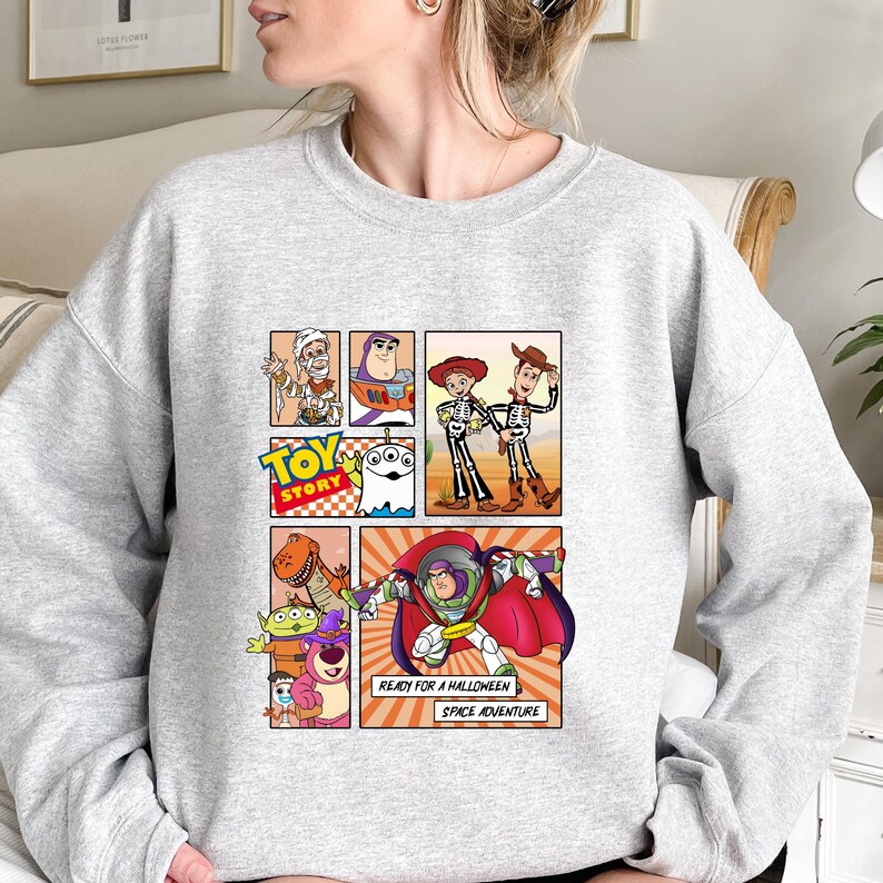 May include: A light gray sweatshirt featuring a comic strip design with characters from the movie "Toy Story." The design includes Buzz Lightyear, Woody, and other characters. Text on the design reads "TOY STORY", "READY FOR A HALLOWEEN SPACE ADVENTURE."