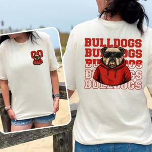 May include: Cream-colored t-shirt featuring a bulldog wearing sunglasses and a red hoodie. The back of the shirt displays the word "BULLDOGS" stacked multiple times. The front has a bulldog graphic and the words "GO Dawgs".