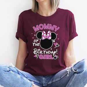 May include: A maroon t-shirt with the text "MOMMY OF THE BIRTHDAY GIRL" in a playful font. The design includes a Minnie Mouse silhouette with a polka dot bow. The shirt is worn by a person wearing blue jeans.