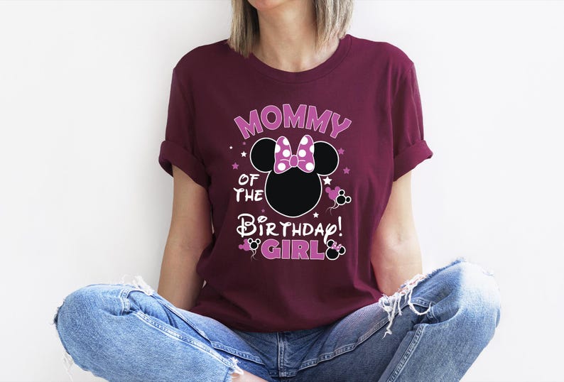 May include: A maroon t-shirt with the text "MOMMY OF THE BIRTHDAY GIRL" in a playful font. The design includes a Minnie Mouse silhouette with a polka dot bow. The shirt is worn by a person wearing blue jeans.