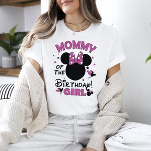 May include: White t-shirt featuring the text "MOMMY OF THE BIRTHDAY GIRL" in pink and black, with a Minnie Mouse head design. The design includes a pink bow and stars. A fun and festive shirt for a birthday celebration.