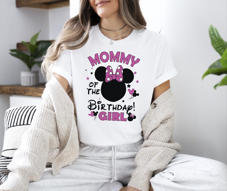May include: White t-shirt featuring the text "MOMMY OF THE BIRTHDAY GIRL" in pink and black, with a Minnie Mouse head design. The design includes a pink bow and stars. A fun and festive shirt for a birthday celebration.