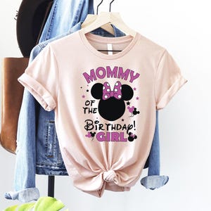 May include: A pale pink t-shirt featuring the text "MOMMY OF THE BIRTHDAY GIRL!" in purple and black. The design includes a black character head silhouette with a pink bow and stars.
