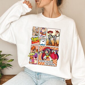 May include: White sweatshirt featuring a comic strip design with characters from the movie "Toy Story." The design includes Buzz Lightyear, Woody, and other characters in Halloween costumes. Text reads "READY FOR A HALLOWEEN SPACE ADVENTURE."