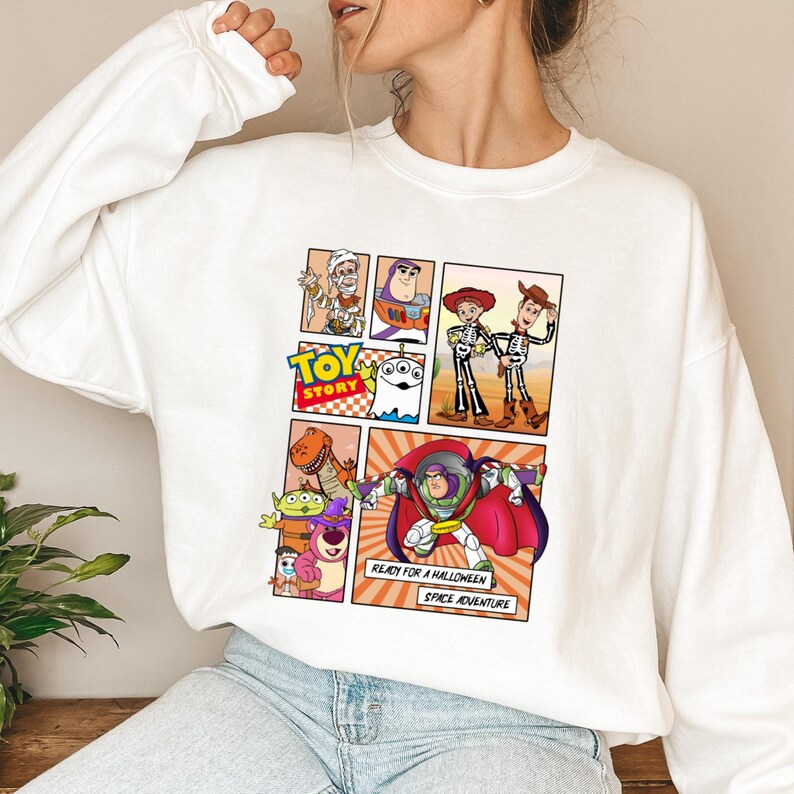 May include: White sweatshirt featuring a comic strip design with characters from the movie "Toy Story." The design includes Buzz Lightyear, Woody, and other characters in Halloween costumes. Text reads "READY FOR A HALLOWEEN SPACE ADVENTURE."