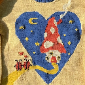Könnte beinhalten: Ein cremefarbener Pullover mit blauem Rippstrick-Ausschnitt. Die Vorderseite zeigt ein blaues Herz mit einem rot-weißen Pilzhaus, gelben Sternen und einem gelben Weg mit roten Marienkäfern.