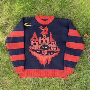 Puede incluir: Jersey azul marino con detalles rojos. El jersey presenta un diseño de castillo rojo en la parte delantera, mangas a rayas rojas y azul marino, y un cuello y puños rojos. Una luna creciente amarilla está en la parte superior izquierda.