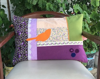 Peut inclure: Un coussin patchwork avec un motif floral violet, vert et orange. Le coussin présente une petite applique d'oiseau orange et trois embellissements de fleurs violettes.