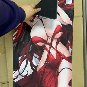 May include: A large rectangular mousepad featuring a stylized anime-style illustration of a woman in a red dress. The mousepad has a black backing and is being held up by a person's hand.