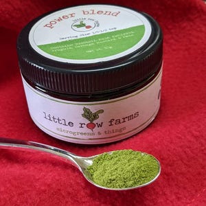 Op de afbeelding: Een potje "power blend" microgreens met een zwart deksel en een wit etiket. Op het etiket staat "little row farms" en "microgreens & things". Een zilveren lepel bevat een portie groen poeder. Het potje bevat 30g product.