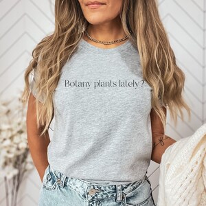 May include: A gray t-shirt with the text "Botany plants lately?" printed on the front. The shirt is being worn by a person with long blonde hair and a gold chain necklace.