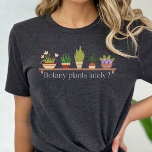 May include: A dark gray t-shirt with a graphic of potted plants and the text "Botany plants lately?" printed on the front.