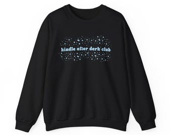 May include: A black long-sleeve sweatshirt featuring a graphic print of light blue stars and crescent moons surrounding the text "kindle after dark club" in a light blue, rounded, lowercase font centered on the chest of the garment.