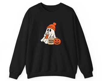 May include: A black crewneck sweatshirt featuring a graphic of a white ghost wearing an orange beanie. The ghost holds an orange drink and a tan tote bag labeled BOOK SHOP, positioned next to an orange jack-o'-lantern with a carved face.