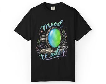 May include: A black t-shirt featuring a graphic print of an oval mirror with a blue and green gradient surface, an open book, a small cup, and leafy sprigs. The text "mood reader" is written in a stylized, colorful script above and below the mirror.
