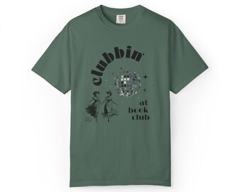 May include: A sage green short-sleeve t-shirt featuring a black graphic print. The design includes a disco ball, two figures in vintage-style dresses, and the text "clubbin' at book club" with small star accents surrounding the central disco ball.
