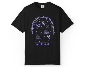 May include: A black short-sleeve t-shirt featuring a light purple graphic inside an arched frame. The design includes a crescent moon, two swords, a crown, an open book, and two dragons, with the text "conquering worlds, dragons, and crowns no big deal."