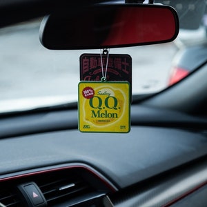May include: A car air freshener with a yellow square design featuring the text "Q.Q. Melon" and "Sadboy". The air freshener is hanging from a car's rearview mirror. The car's dashboard is black with red trim.