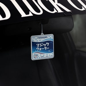 Puede incluir: Un ambientador de coche cuadrado blanco con texto azul y blanco que dice "ミチノクサイダー" y "マジックウォーター".