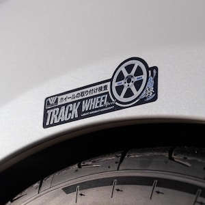 May include: A close-up of a silver car with a black tire and a sticker that reads "TRACK WHEEL LIGHT WEIGHT CONCEPT" in Japanese and English. The sticker also features a stylized image of a wheel.