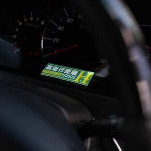 Könnte beinhalten: Ein grüner und gelber Aufkleber mit dem Text "High Mileage Club 100000" und dem japanischen Text "高走行距離" auf einem schwarzen Armaturenbrett eines Autos. Der Aufkleber befindet sich neben dem Tachometer des Autos.