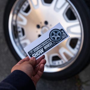 May include: A white sticker with black text and a small image of a wheel. The text reads "Show Wheel" and "Personalized Fitment". The sticker is held in front of a silver car wheel.