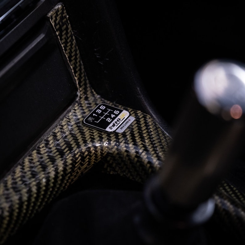 Puede incluir: Primer plano de un entorno de palanca de cambios de fibra de carbono negro y dorado con una pegatina en blanco y negro que dice "5 Speed", "1 3 5", "2 4 6" y "M15B15".