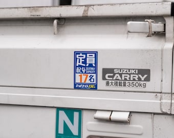 Może przedstawiać: Biały samochód dostawczy Suzuki Carry z niebiesko-pomarańczową naklejką z napisem "Pojemność miejsc siedzących 17" i szarą naklejką z napisem "Suzuki Carry Maksymalny ładunek 350 kg".