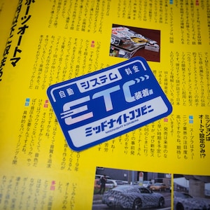 Pode incluir: Etiqueta azul e branca com o texto "Sistema autom&aacute;tico de ped&aacute;gio ETC Midnight Konbini" em japon&ecirc;s. A etiqueta &eacute; projetada para parecer um passe de ped&aacute;gio.