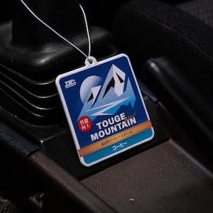 Op de afbeelding: Een blauwe en witte autoluchtverfrisser met de tekst "Touge Mountain Blend" en "Coffee" in het Japans. De luchtverfrisser is gevormd als een berg en heeft een rode sticker met de tekst "性能向上" in het Japans.