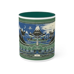 May include: A white ceramic mug with a green rim and a colorful illustration of the cover of the book "The Hobbit" by J.R.R. Tolkien. The illustration features a mountain range, a river, and a dragon.