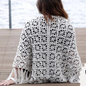 May include: A cream-colored, open-weave crochet shawl with a floral pattern and fringe trim. The shawl is draped over the shoulders. The text "Häkelanleitung Stola MIRABEL" is visible.