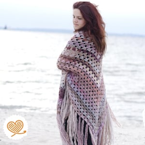 May include: A woman wearing a crocheted shawl in shades of pink, white, and brown. The shawl has a fringe border. The text on the image reads "Stola häkeln AVA Anleitung in deutscher Sprache". A German flag is also visible.