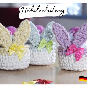 Può includere: Tre cestini pasquali all'uncinetto a forma di coniglio con fiocchi colorati. I cestini sono realizzati in filato bianco e hanno orecchie di coniglio gialle, viola e verdi. I cestini sono pieni di uova di cioccolato.