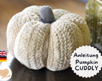 May include: A cream-colored crocheted pumpkin with a textured, ribbed surface and a small, dark gray crocheted stem at the top. A white scalloped label on the right side features the text 'Anleitung Pumpkin CUDDLY' in dark gray lettering.